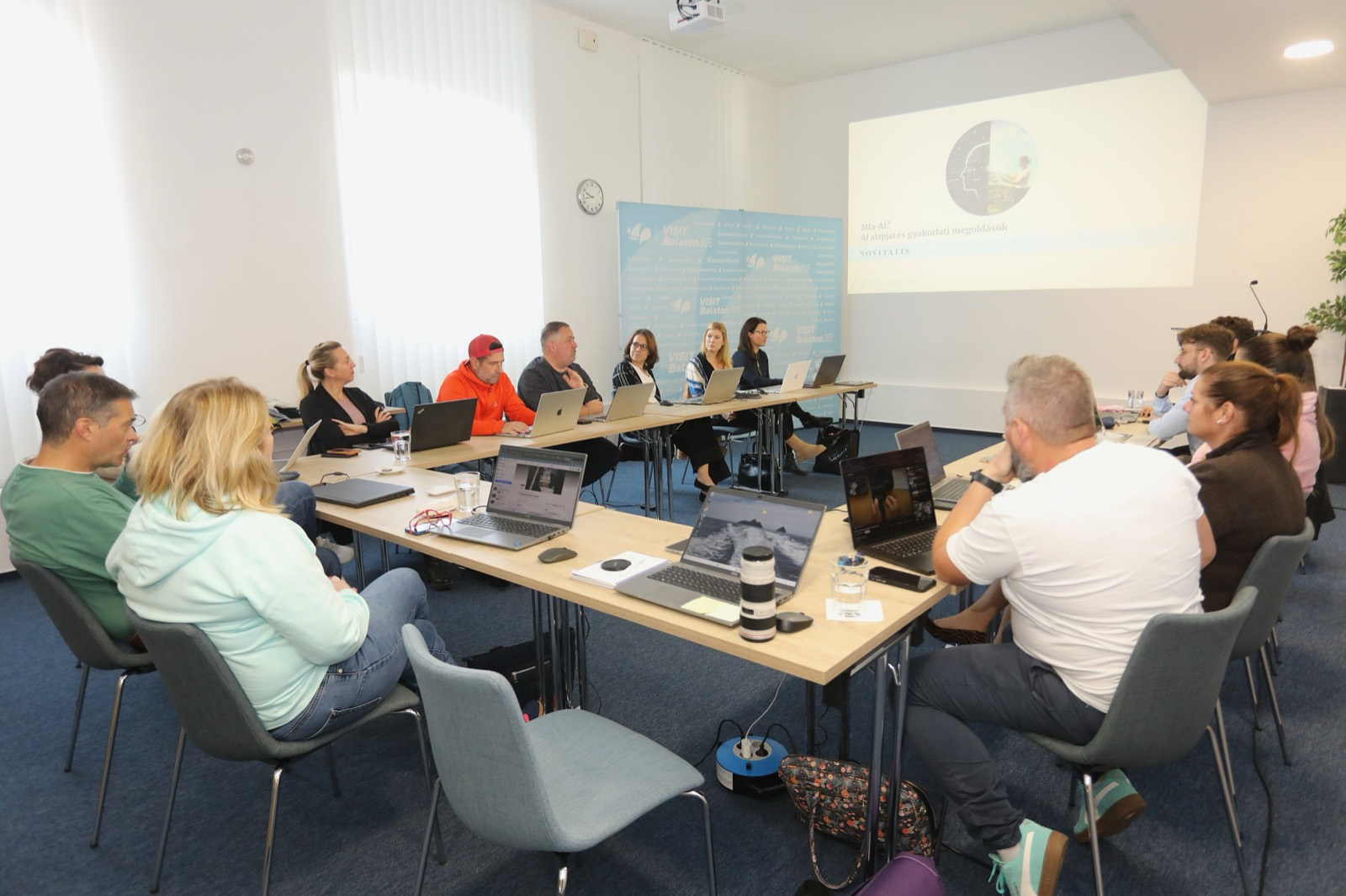 01: AI training in the VisitBalaton365 training room in Keszthely; participants working on laptops, the AI slide on the screen, the map of Lake Balaton on the wall. 02: VisitBalaton365 DMO staff at the U-shaped tables during the AI training, in front of the Lake Balaton map wall. 03: Participants practicing AI tools on laptops during the training. 04: The full group of the training room at work. 05: DMO staff attentively following the AI training demo.