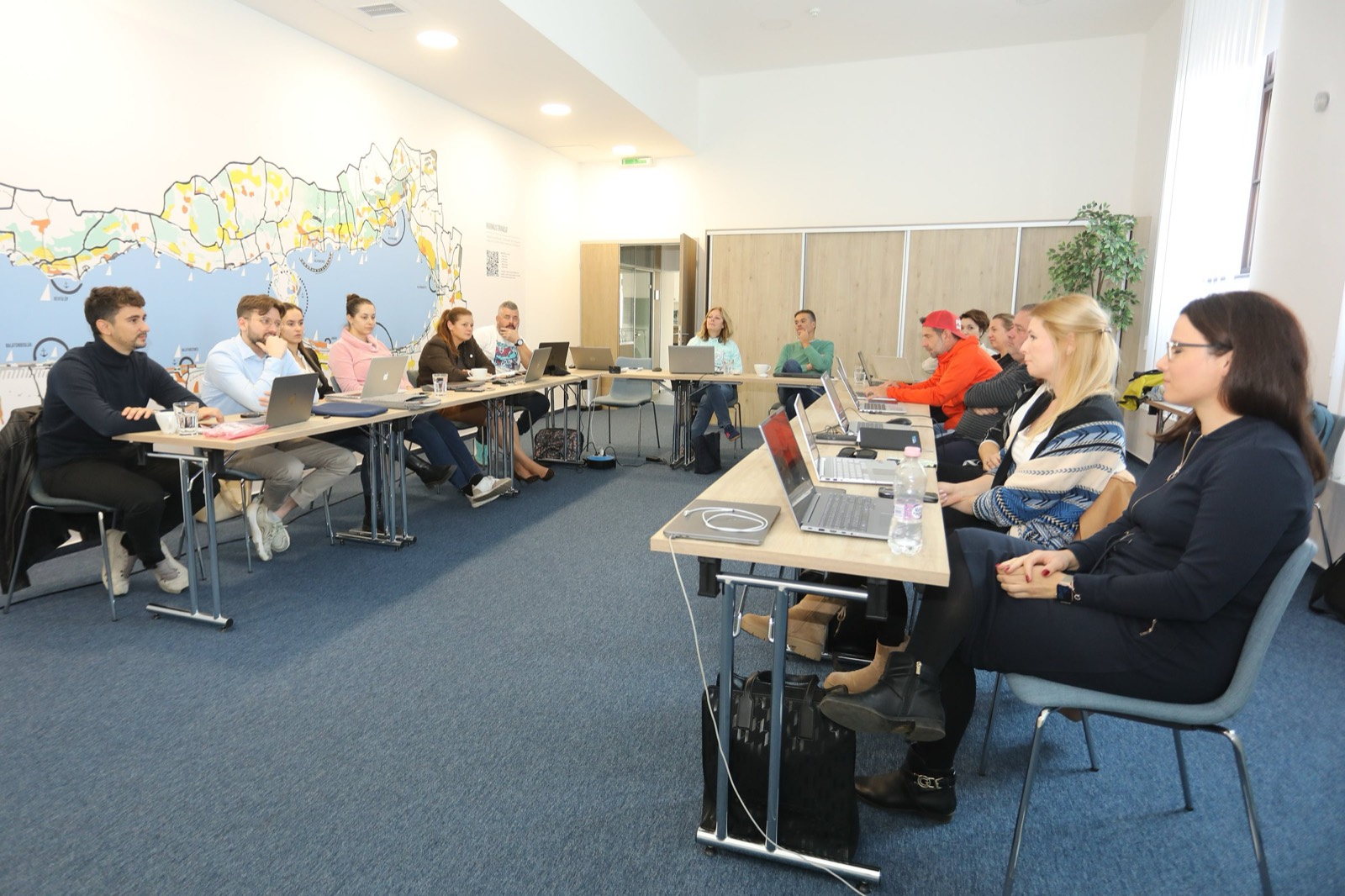 01: AI training in the VisitBalaton365 training room in Keszthely; participants working on laptops, the AI slide on the screen, the map of Lake Balaton on the wall. 02: VisitBalaton365 DMO staff at the U-shaped tables during the AI training, in front of the Lake Balaton map wall. 03: Participants practicing AI tools on laptops during the training. 04: The full group of the training room at work. 05: DMO staff attentively following the AI training demo.