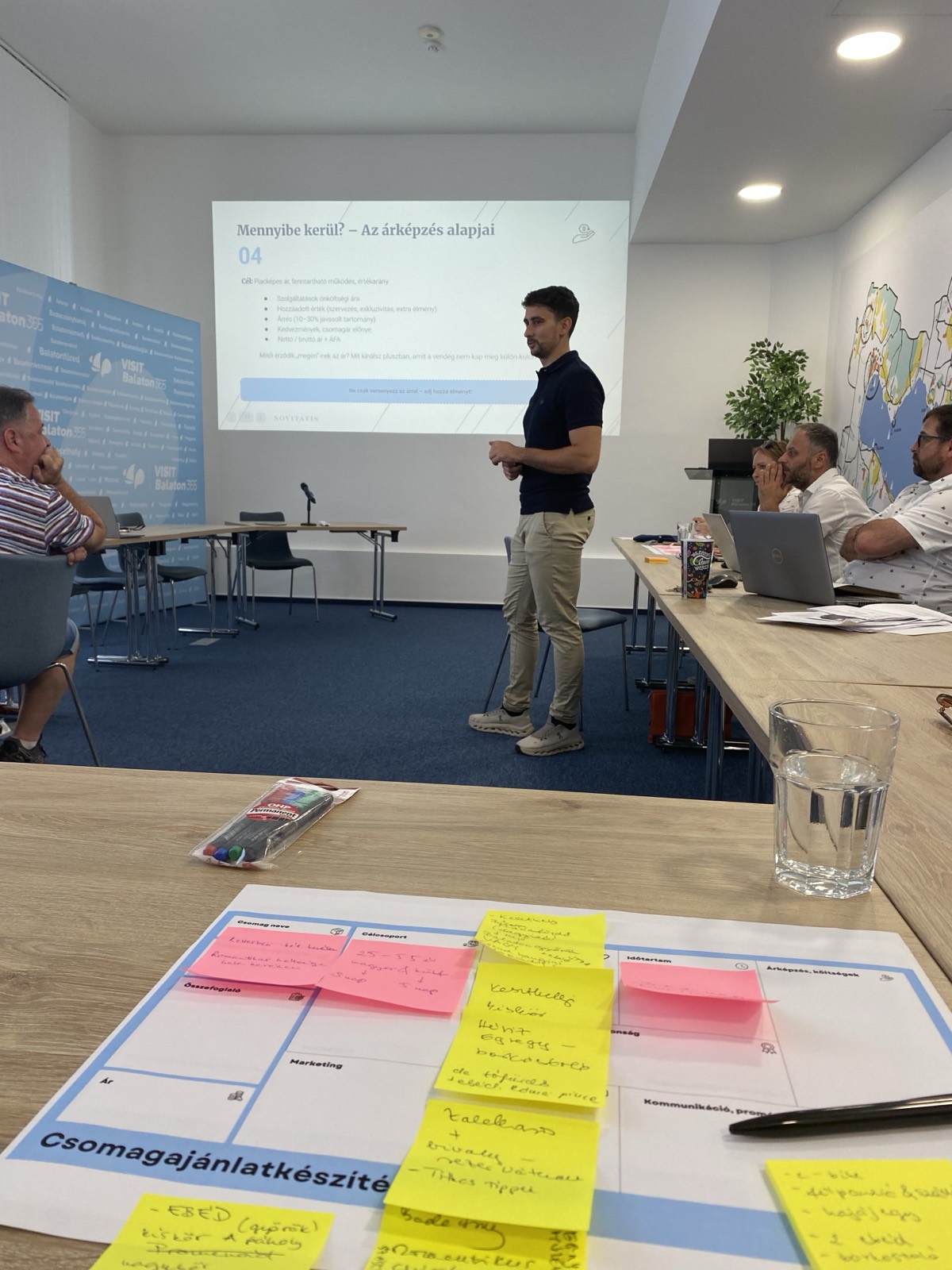 01: Adam Schmutz giving a talk on package pricing at VisitBalaton365's Keszthely training center; in the foreground the “Package creation” canvas with participants' notes. 02: The Keszthely training room with the Lake Balaton map on the wall; participants at the two-day workshop, the title slide on the screen. 03: Small-group work next to the VisitBalaton365 banner and the projected canvas template. 04: Participants following a package example on the projector. 05: The Keszthely training room from above, participants at the U-shaped tables in front of the Lake Balaton map wall. 06: A small group working together on the package canvas. 07: Participants working over the package-planning template at the long table. 08: A lively discussion in a four-person group. 09: Workshop participants in a small-group consultation with notes and a canvas. 10: Participants at the training room table in front of the Lake Balaton map.