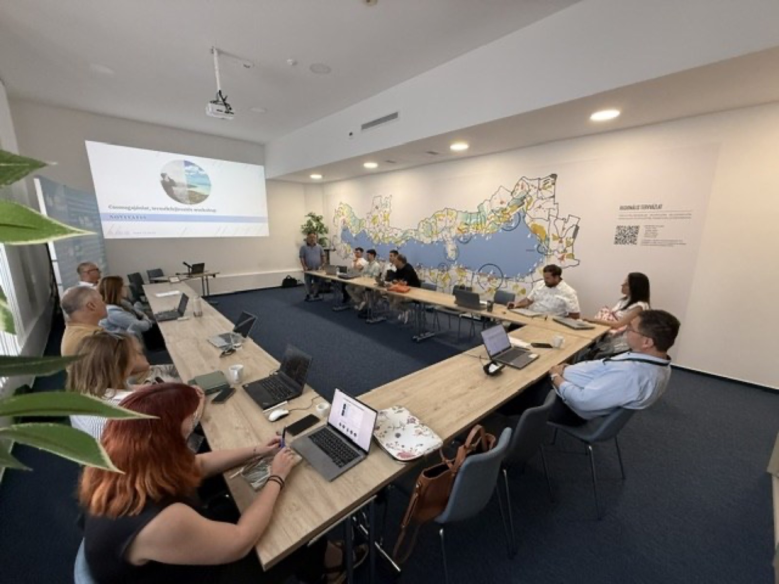 01: Adam Schmutz giving a talk on package pricing at VisitBalaton365's Keszthely training center; in the foreground the “Package creation” canvas with participants' notes. 02: The Keszthely training room with the Lake Balaton map on the wall; participants at the two-day workshop, the title slide on the screen. 03: Small-group work next to the VisitBalaton365 banner and the projected canvas template. 04: Participants following a package example on the projector. 05: The Keszthely training room from above, participants at the U-shaped tables in front of the Lake Balaton map wall. 06: A small group working together on the package canvas. 07: Participants working over the package-planning template at the long table. 08: A lively discussion in a four-person group. 09: Workshop participants in a small-group consultation with notes and a canvas. 10: Participants at the training room table in front of the Lake Balaton map.