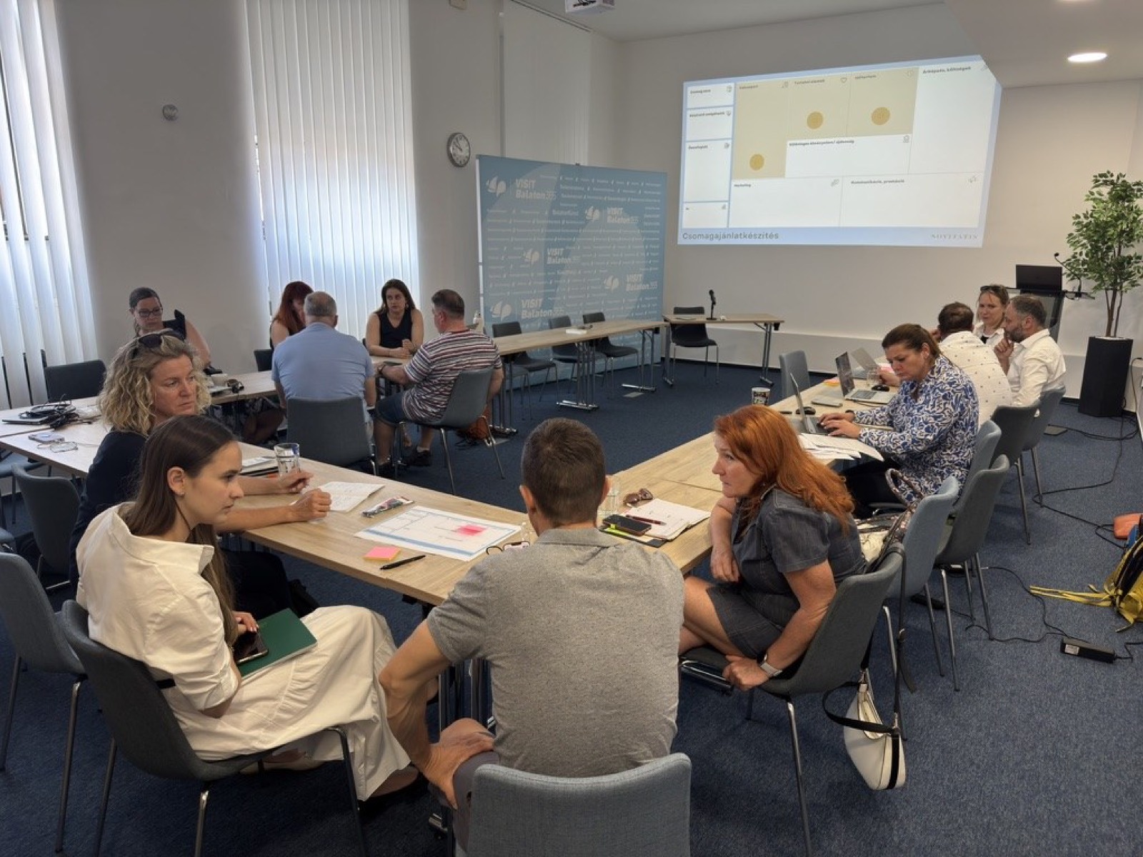 01: Adam Schmutz giving a talk on package pricing at VisitBalaton365's Keszthely training center; in the foreground the “Package creation” canvas with participants' notes. 02: The Keszthely training room with the Lake Balaton map on the wall; participants at the two-day workshop, the title slide on the screen. 03: Small-group work next to the VisitBalaton365 banner and the projected canvas template. 04: Participants following a package example on the projector. 05: The Keszthely training room from above, participants at the U-shaped tables in front of the Lake Balaton map wall. 06: A small group working together on the package canvas. 07: Participants working over the package-planning template at the long table. 08: A lively discussion in a four-person group. 09: Workshop participants in a small-group consultation with notes and a canvas. 10: Participants at the training room table in front of the Lake Balaton map.