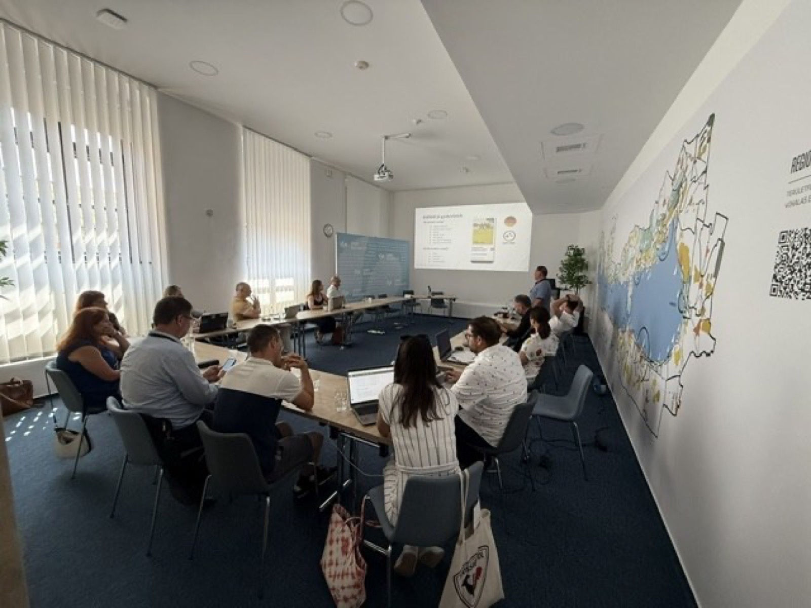 01: Adam Schmutz giving a talk on package pricing at VisitBalaton365's Keszthely training center; in the foreground the “Package creation” canvas with participants' notes. 02: The Keszthely training room with the Lake Balaton map on the wall; participants at the two-day workshop, the title slide on the screen. 03: Small-group work next to the VisitBalaton365 banner and the projected canvas template. 04: Participants following a package example on the projector. 05: The Keszthely training room from above, participants at the U-shaped tables in front of the Lake Balaton map wall. 06: A small group working together on the package canvas. 07: Participants working over the package-planning template at the long table. 08: A lively discussion in a four-person group. 09: Workshop participants in a small-group consultation with notes and a canvas. 10: Participants at the training room table in front of the Lake Balaton map.