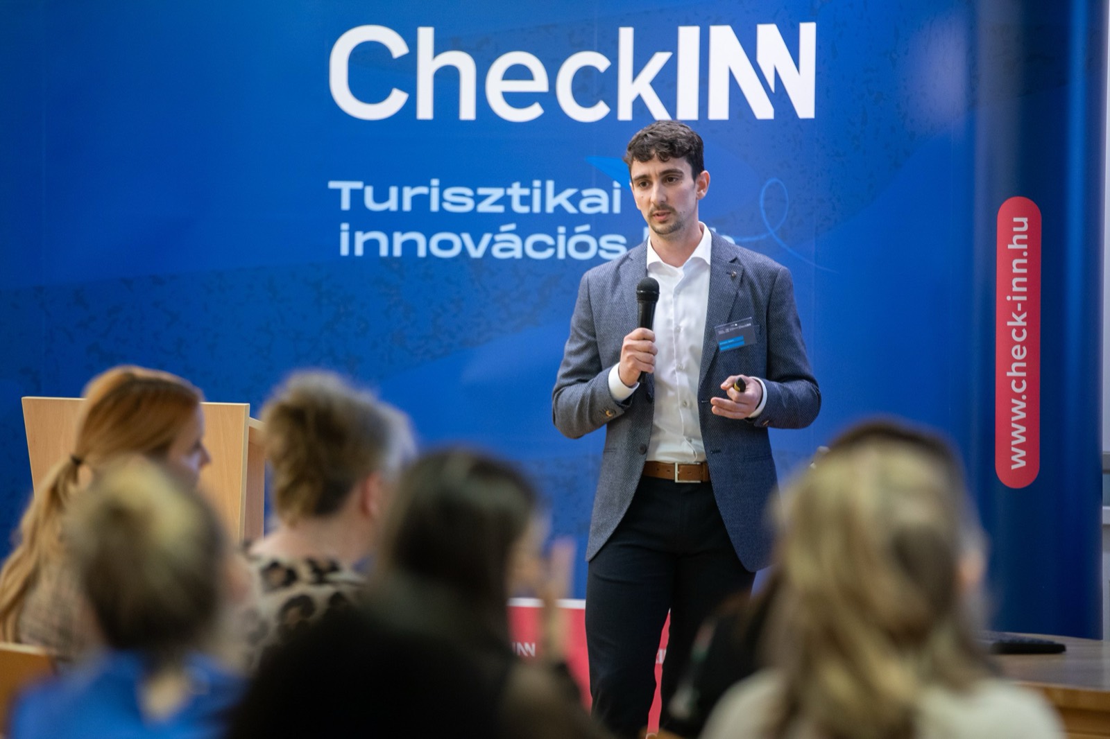 01: Adam Schmutz speaking with a microphone in front of the CheckINN banner, with the audience in the foreground. 02: Adam Schmutz mid-talk, the CheckINN Tourism Innovation Hub banner in the background. 03: Adam Schmutz presenting to the workshop audience in front of the CheckINN backdrop. 04: The full lecture hall with the audience; Adam Schmutz on stage by the CheckINN banner. 05: Round-table discussion: Adam Schmutz and the moderator in front of the METU backdrop. 06: The participants of the workshop's round-table discussion in front of the Budapest Metropolitan University banner. 07: Adam Schmutz during a professional conversation in a break of the event.